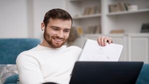 Pessoa sentada em sofá estudando com caderno e laptop, usando fones de ouvido