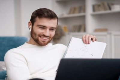Pessoa sentada em sofá estudando com caderno e laptop, usando fones de ouvido